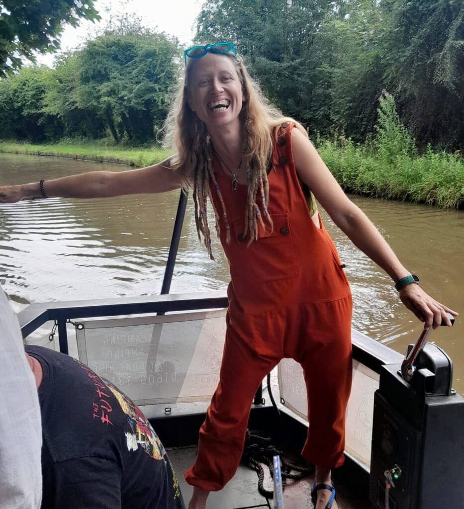 Missy captain-ing a barge up a canal in England, wearing orange dungarees and sporting messy hair. They have a huge smile on their face, exuding joy and adventure as they navigate the peaceful waterway."